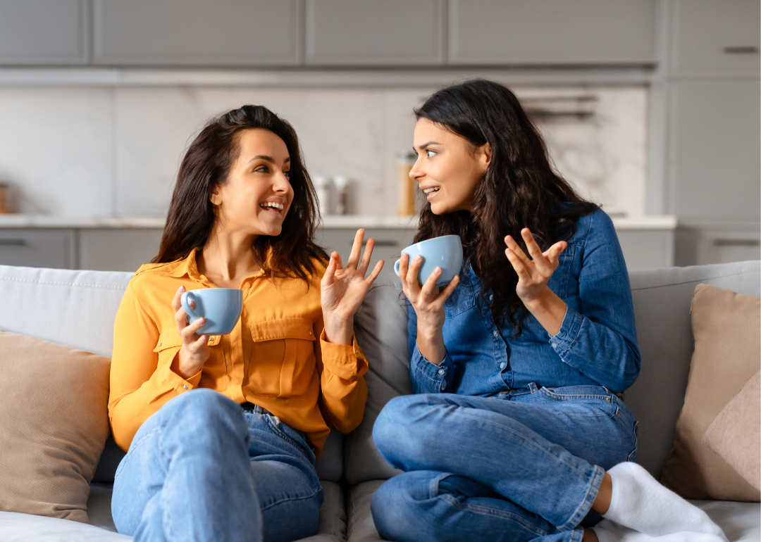 Two young women share stories over tea on a beige couch at home