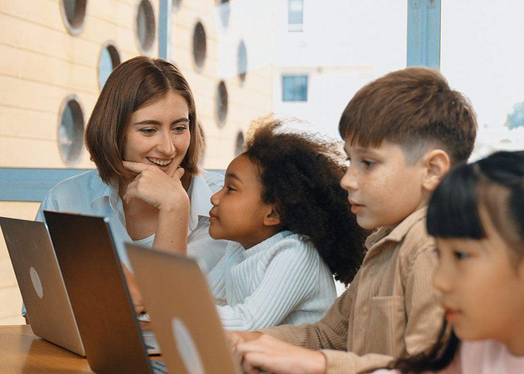 A female teacher looking at three young students while they all use laptops.