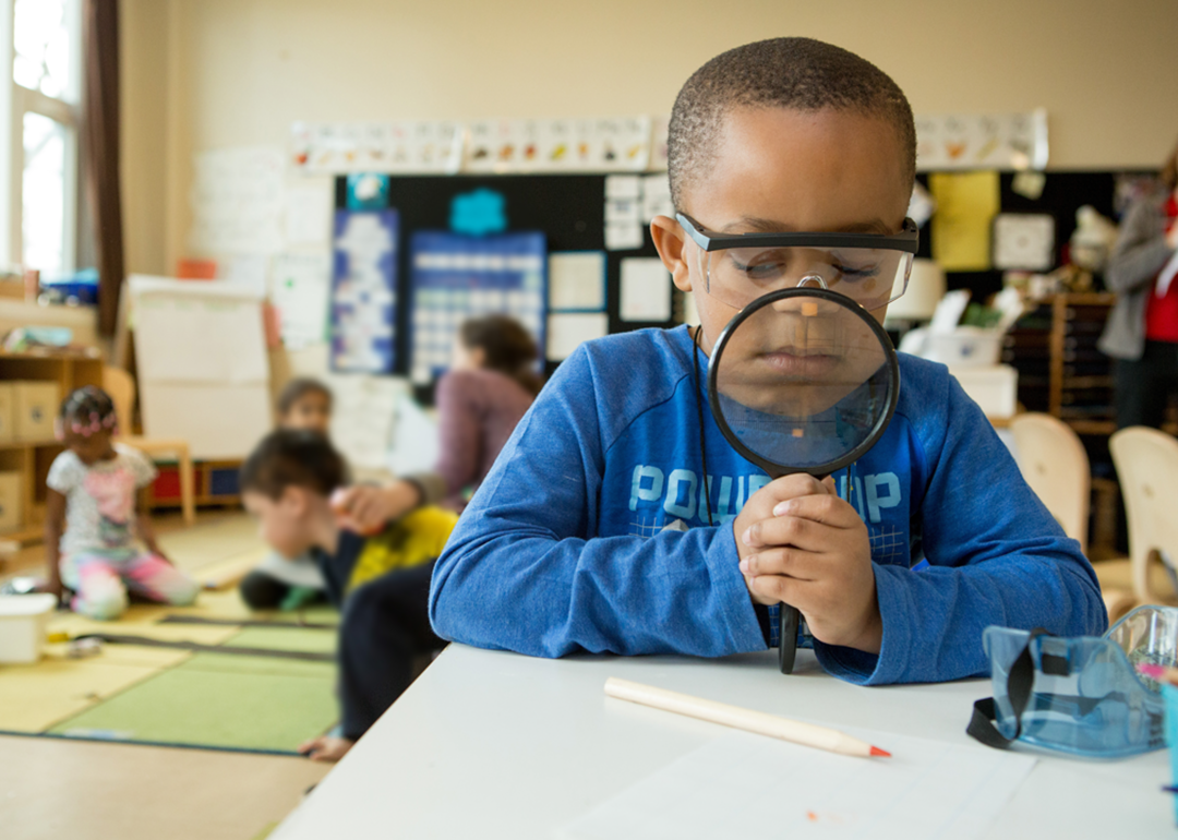 A child in a classroom looks through a magnifying glass down at learning materials on his desk. The magnifying glass makes the bottom of his face appear larger.