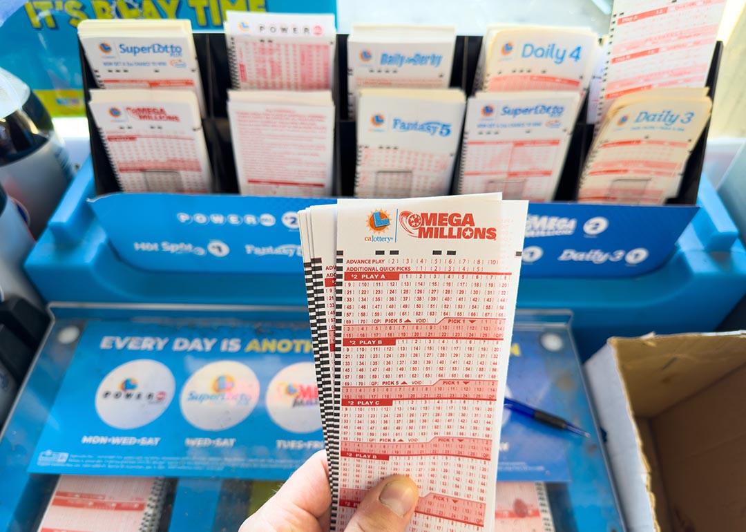 View of left hand holding Mega Millions tickets in front of a blue lottery kiosk.