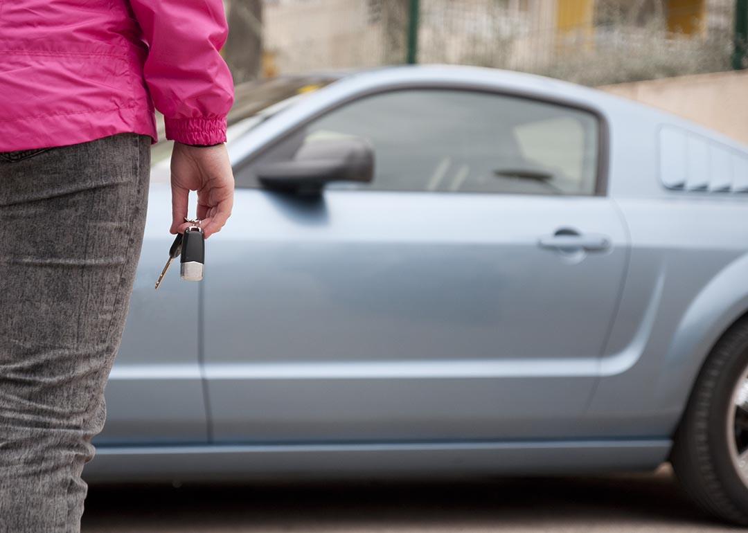 Person in dark pink shirt and black jeans holding car keys seen from behind, standing in front of used light blue car. 