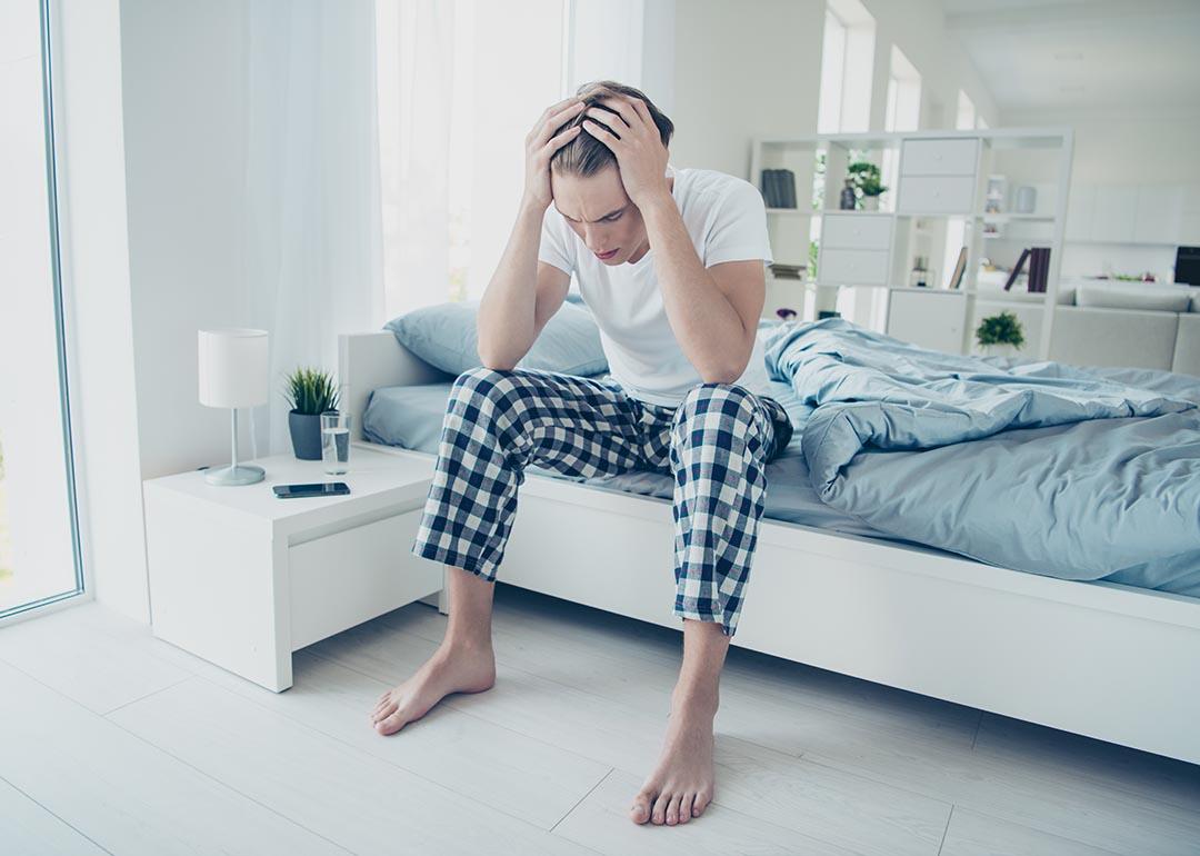 Young adult male sitting on a bed with both hands holding his head seemingly having a headache.