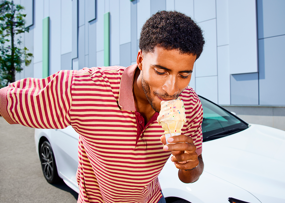 Man eats an ice cream cone in front of a white car in the background.