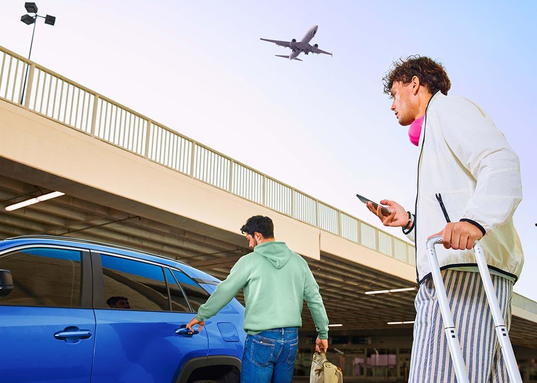 Two travelers head to a blue car with luggage while a plane flies over head.