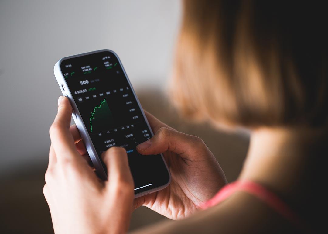 Woman seen from behind, holding a phone investment app showing index growth chart.