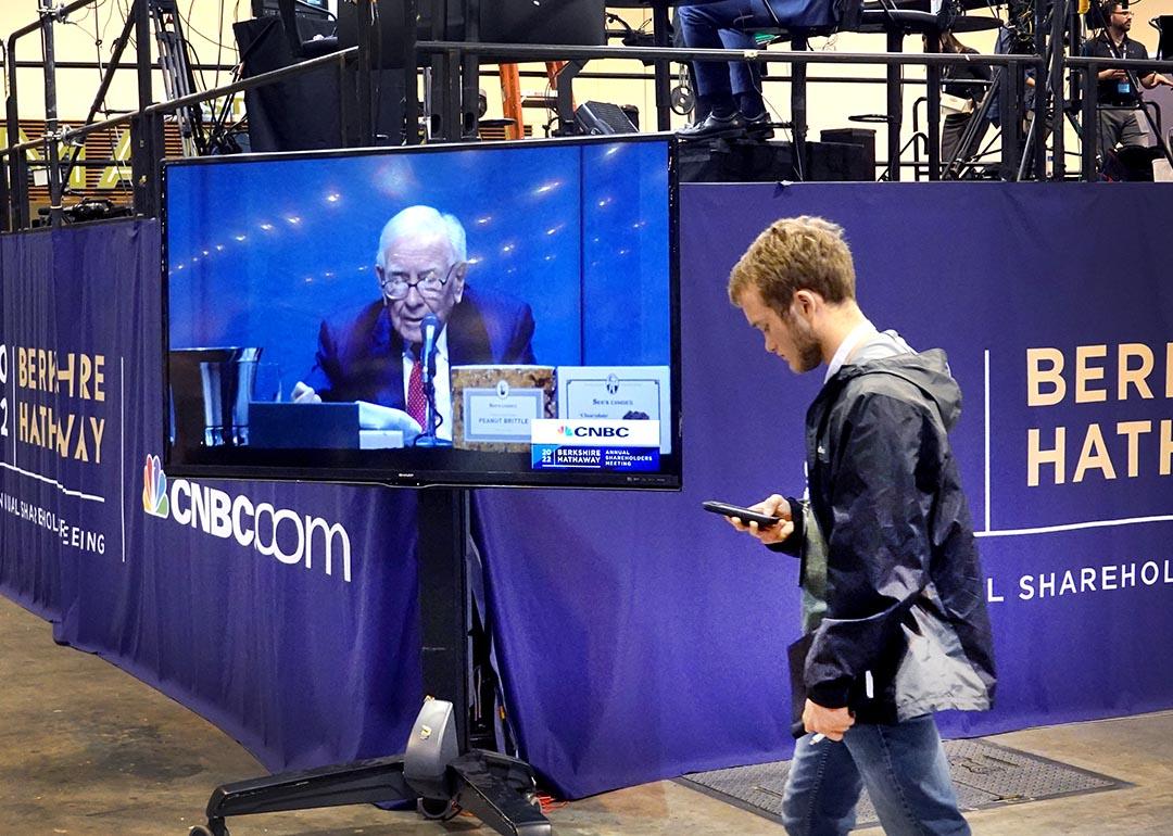 Warren Buffett appears on a TV screen at the Berkshire Hathaway sharesholders meeting, while a bystander walks past.