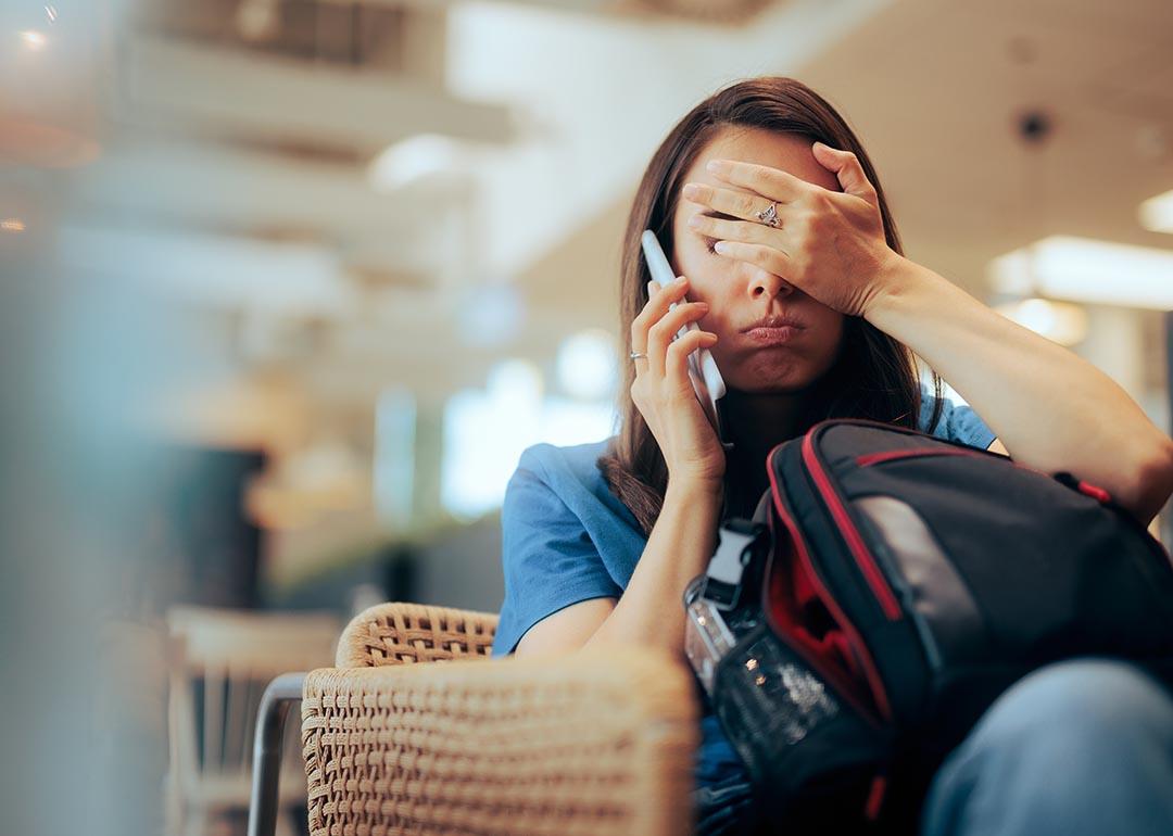 Person on the phone at airport gate, showing signs of frustration covering face with hand while sitting with luggage.