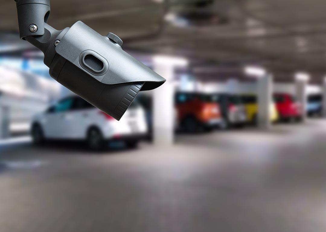 Close up of a security camera in focus inside an indoor parking garage, with blurred cars in the background.