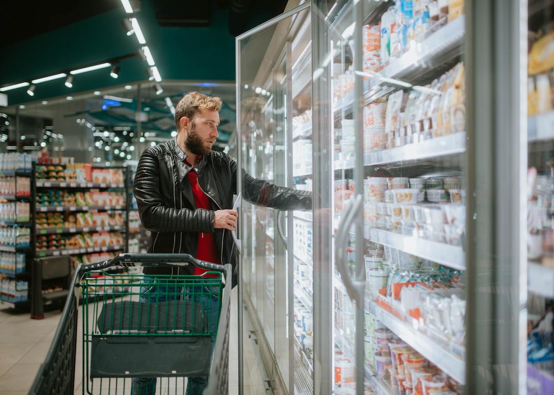 A grocery shopper in a leather jacket pushes a cart through the supermarket and takes food from the fridge.