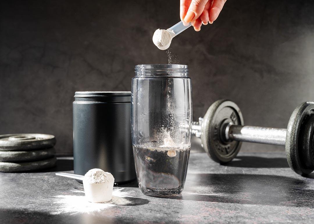 Creatine powder in a measuring scoop being dropped into a clear plastic water bottle, surrounded by workout accessories.