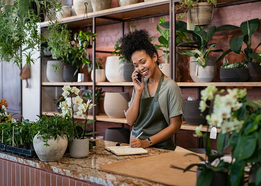 A female owner of a small botanical shop smiling while on the phone taking an order.