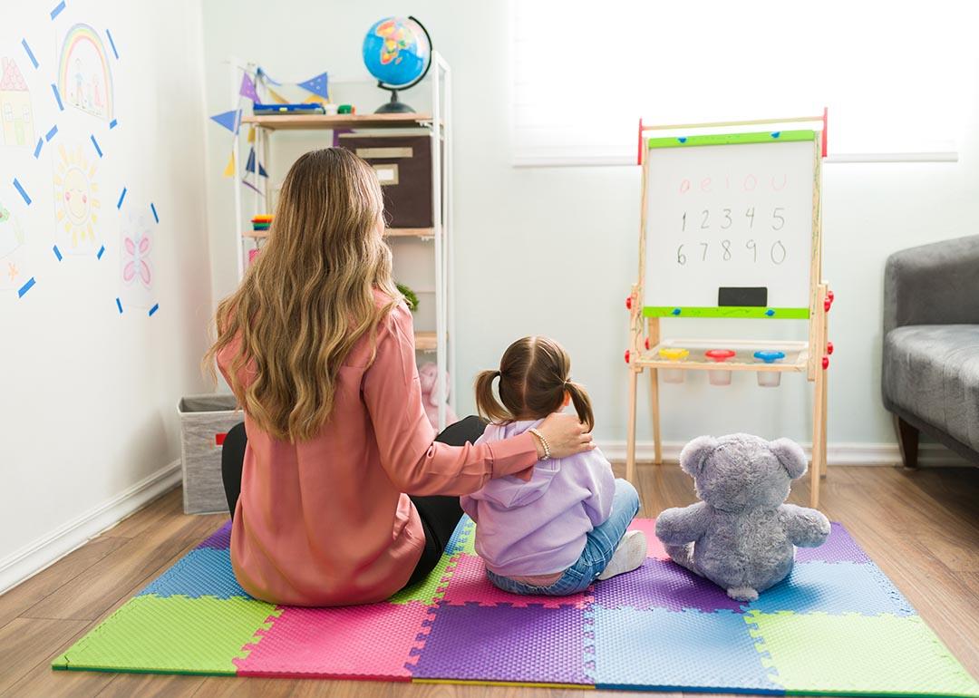 Preschooler in classroom with adult figure sitting on mat as seen from the back, next to a plush toy. 