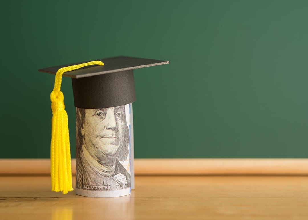 Concept of student debt illustrated by a rolled up dollar bill wears graduation cap with a chalkboard background in classroom.