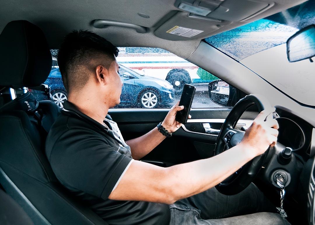 Distracted driver looks at cell phone in one hand with other hand on steering wheel while other cars can be seen through driver window. 