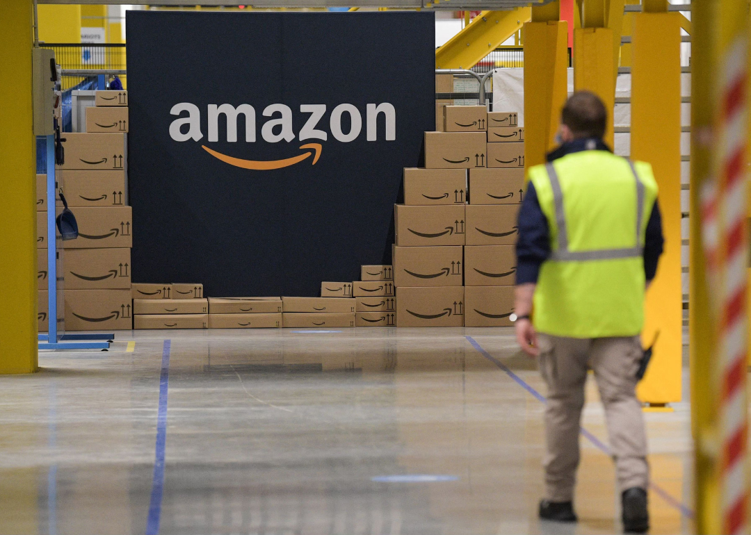 An Amazon warehouse employee walking towards a pile of Amazon-branded boxes and a large Amazon logo sign