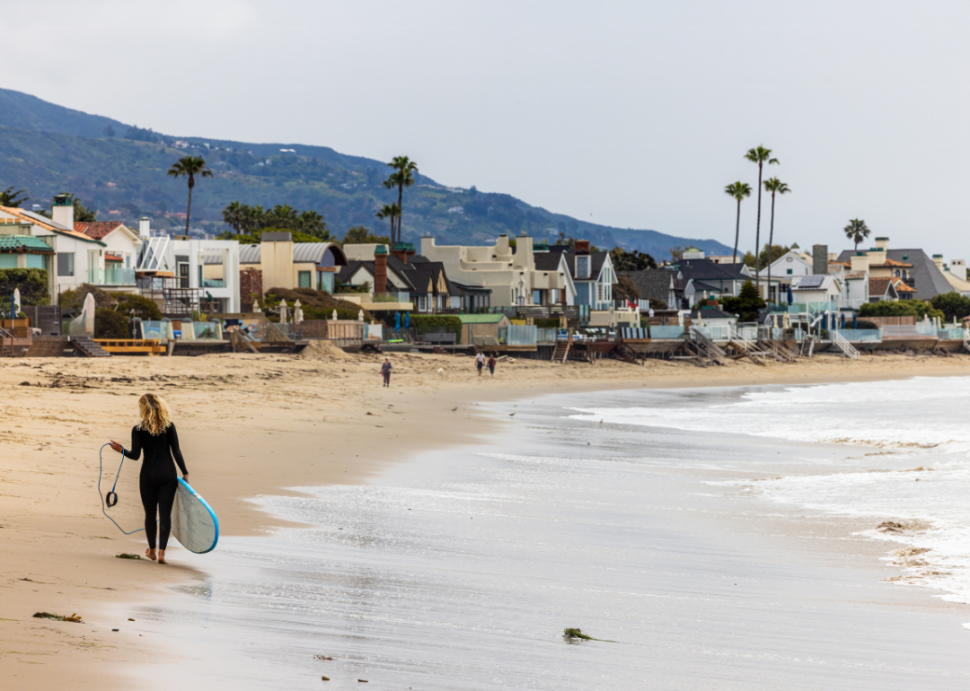 A surfer in a wet suit walking along the beach in Malibu, California carrying a surfboard.