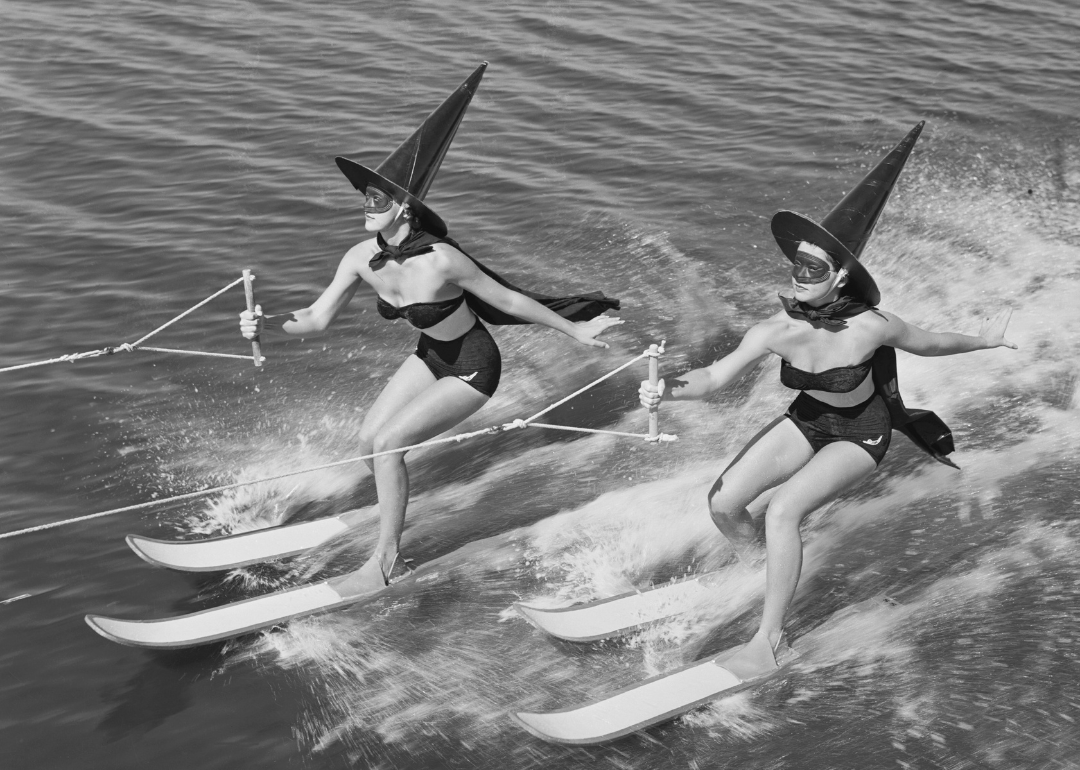 Two women dressed in black swimsuits and witch hats on jet skis