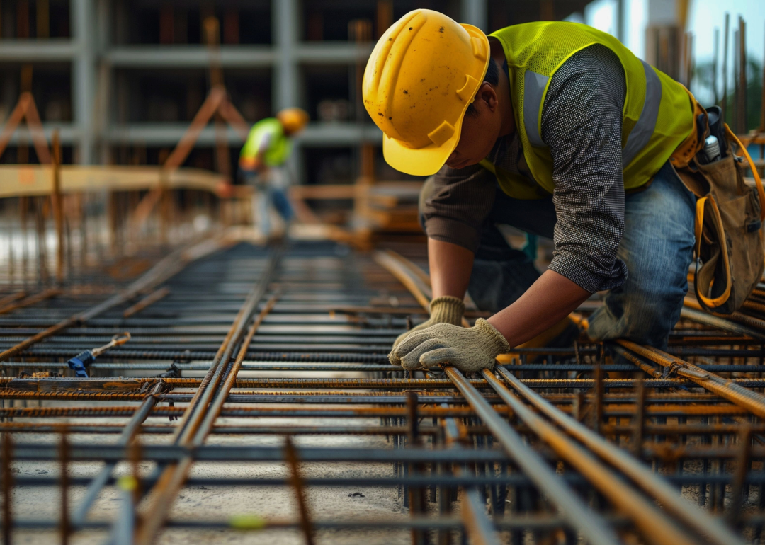 A construction worker wearing yellow hard hat kneeling down on metal rods at a construction site, focused on his task.