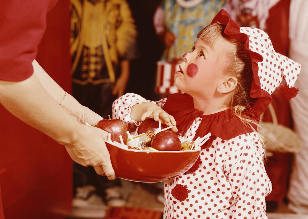 A child in a clown costume choosing a treat from a bowl of apples and candy during Halloween in the 1960s.