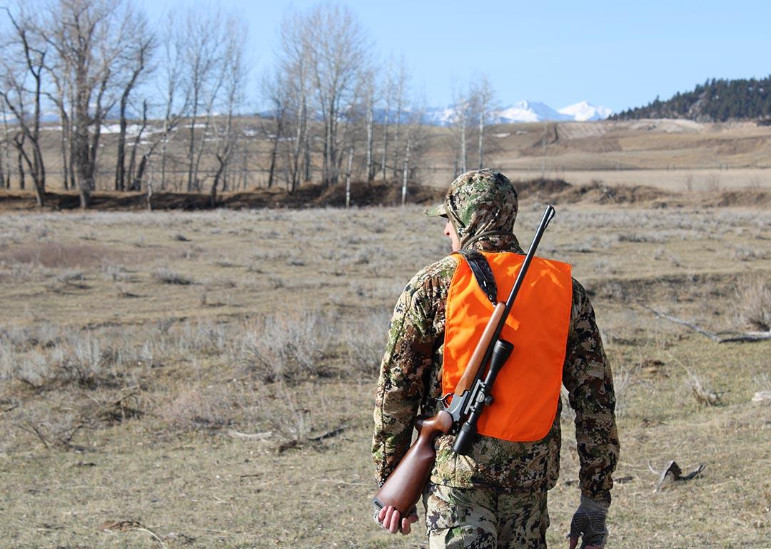 Rear view of a hunter carrying a rifle and wearing high-visibility vest walking toward open field.