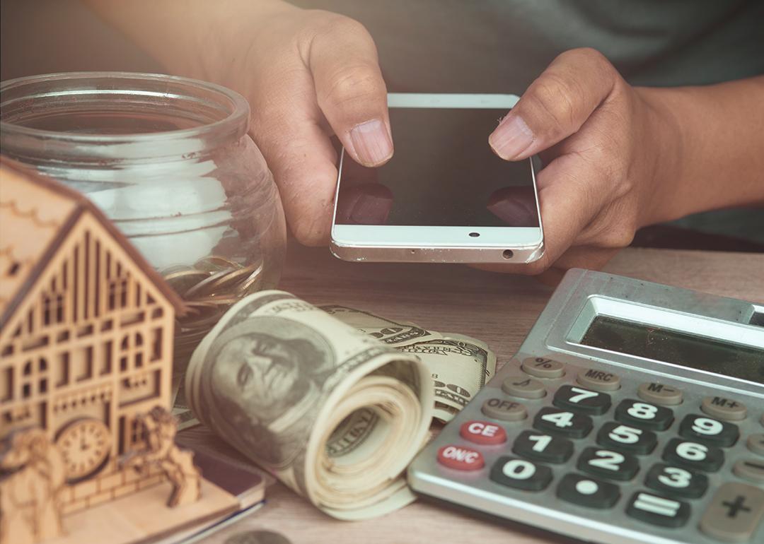 Person holding a phone with money, a figure of a house and savings around to represent passive income.