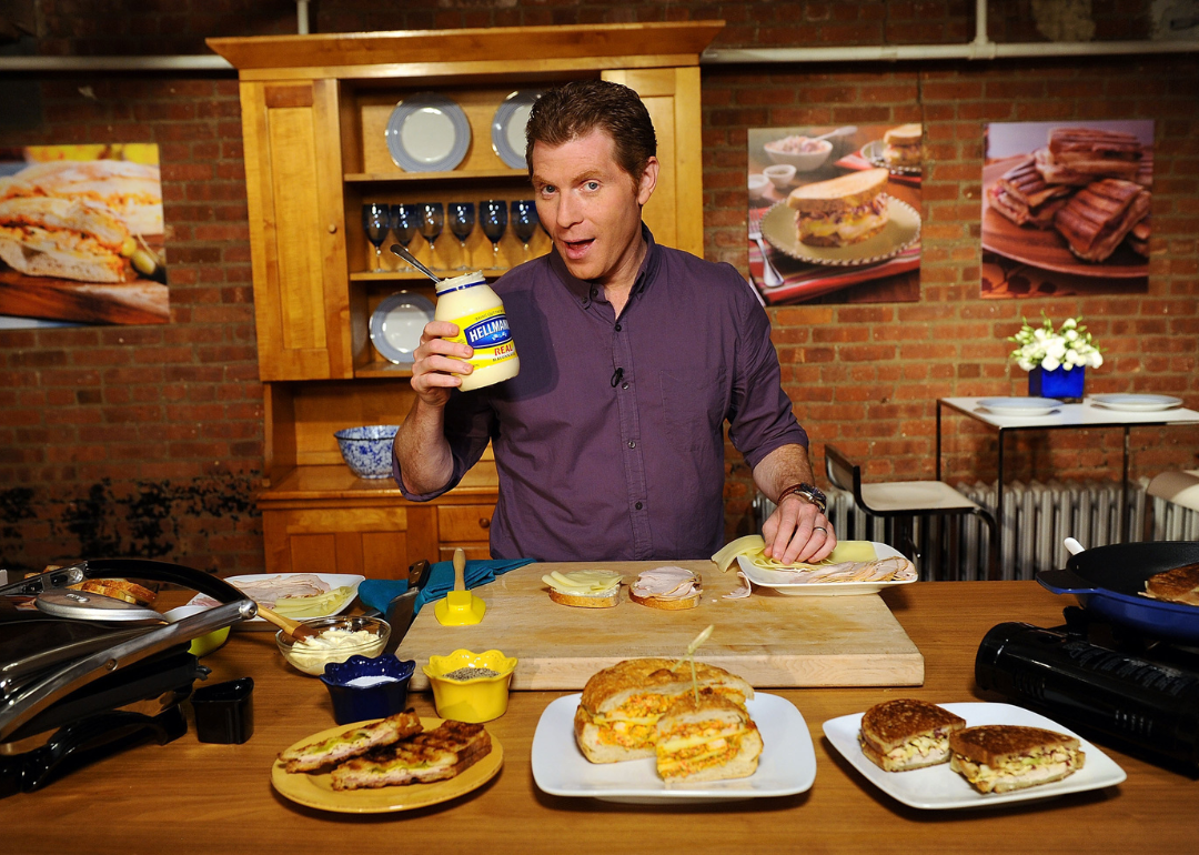 Chef Bobby Flay holding up a jar of mayonnaise in front of a countertop with plates of food, circa 2011.