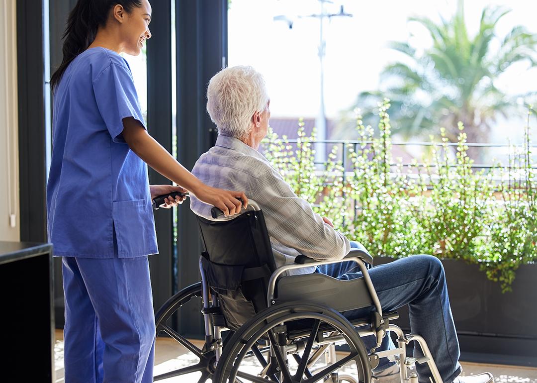 A senior man in a wheelchair looking out a balcony and assisted by a nurse.