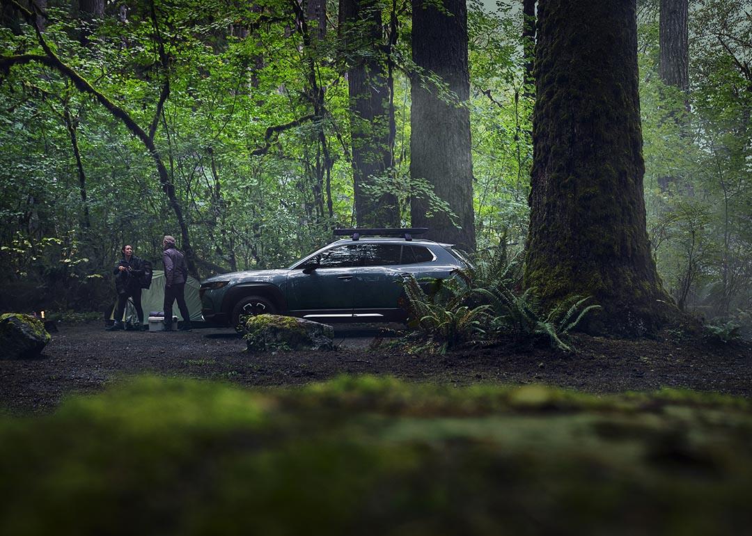 Dark gray Mazda car parked in a forest where two adults are setting up a tent.