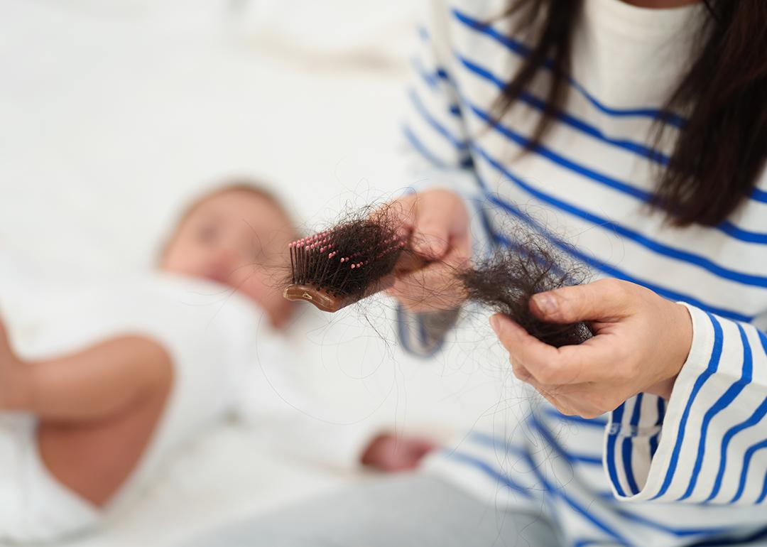 A mother looks at a brush with excessive hair fall and her newborn baby is beside her.