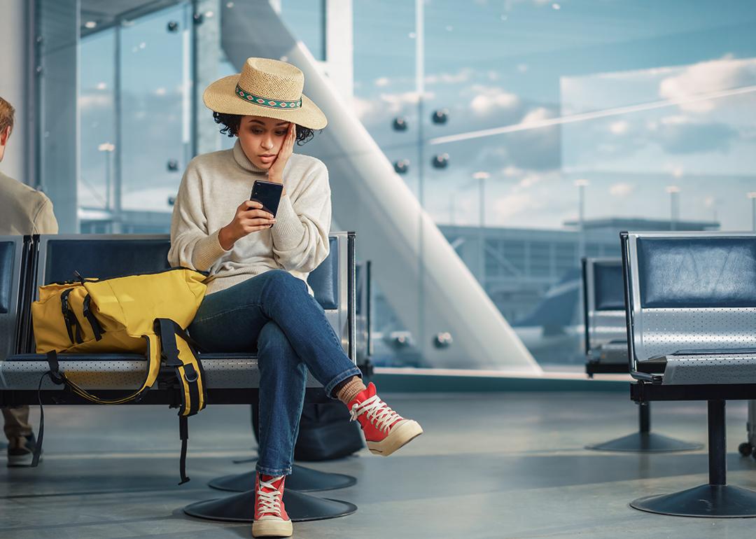 A woman in an airport is checking flight updates in phone while looking worried.