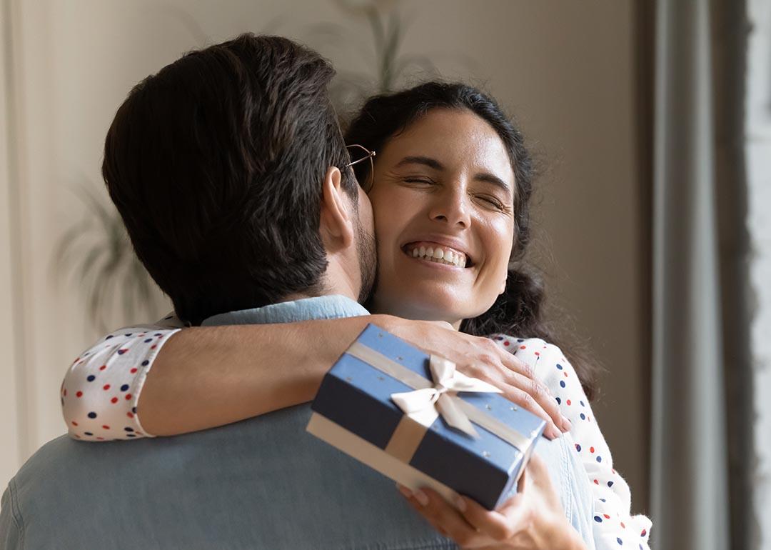 Woman smiling with eyes closed hugging a man whose back is to the camera, she is holding a small blue box tied with a bow.