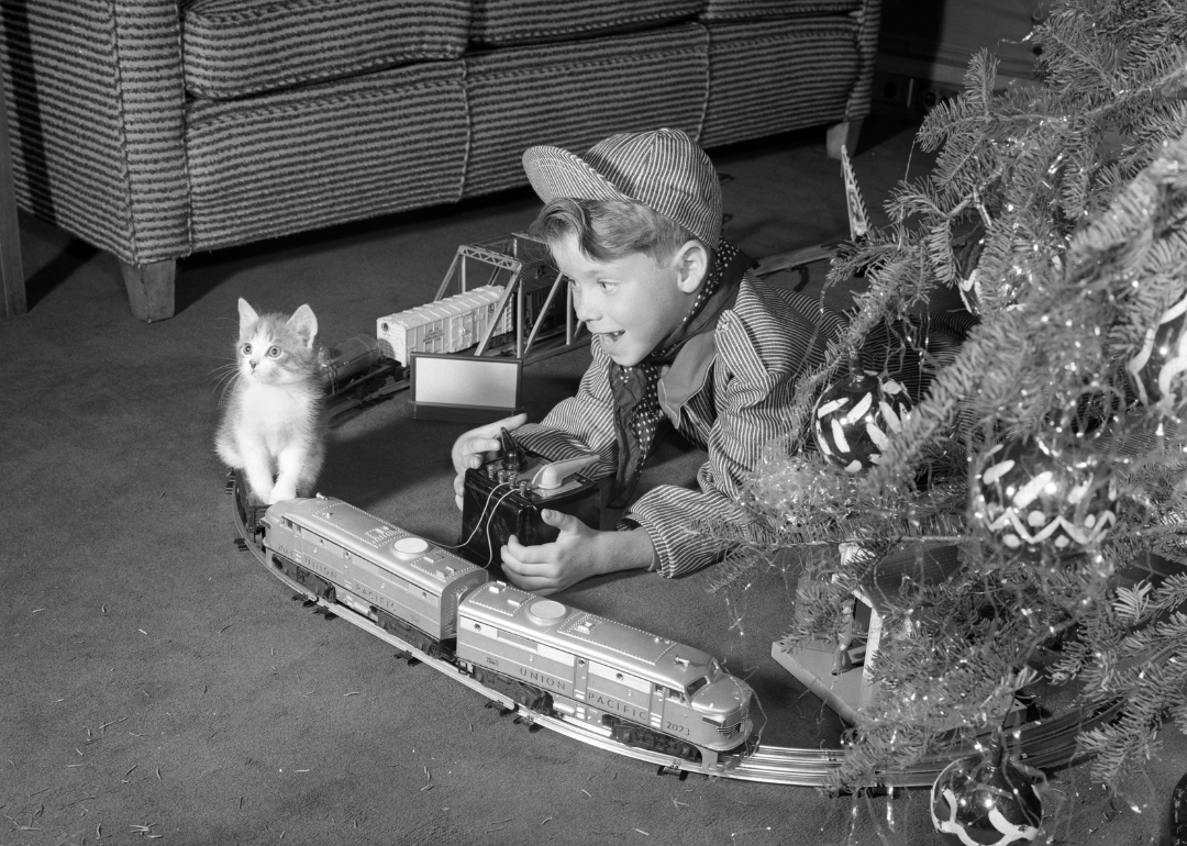 Young boy dressed as an engineer and his kitten play with a train set next to a Christmas tree