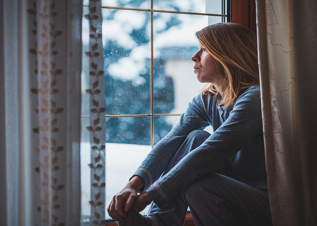 A woman sadly sitting by and looking out a window during winter.