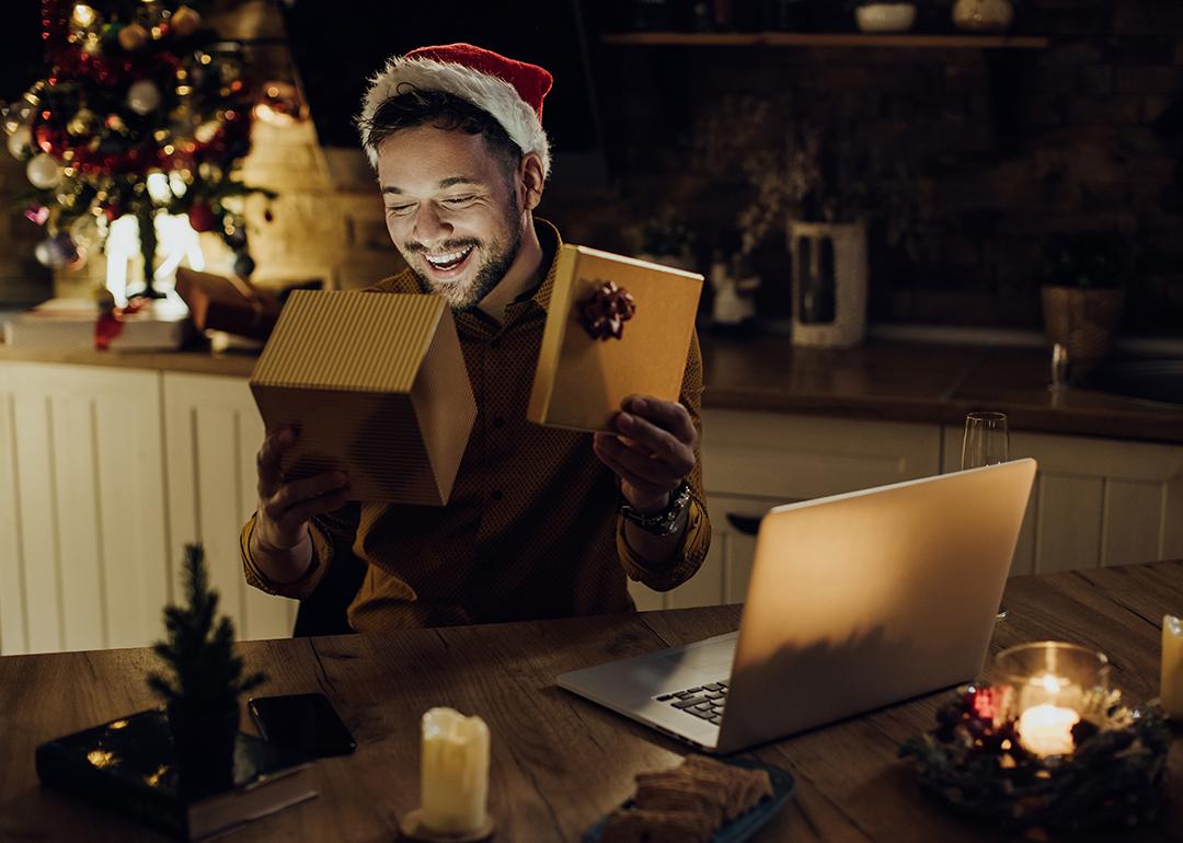 A young man happily opening Christmas presents while in a video call with someone on a laptop.