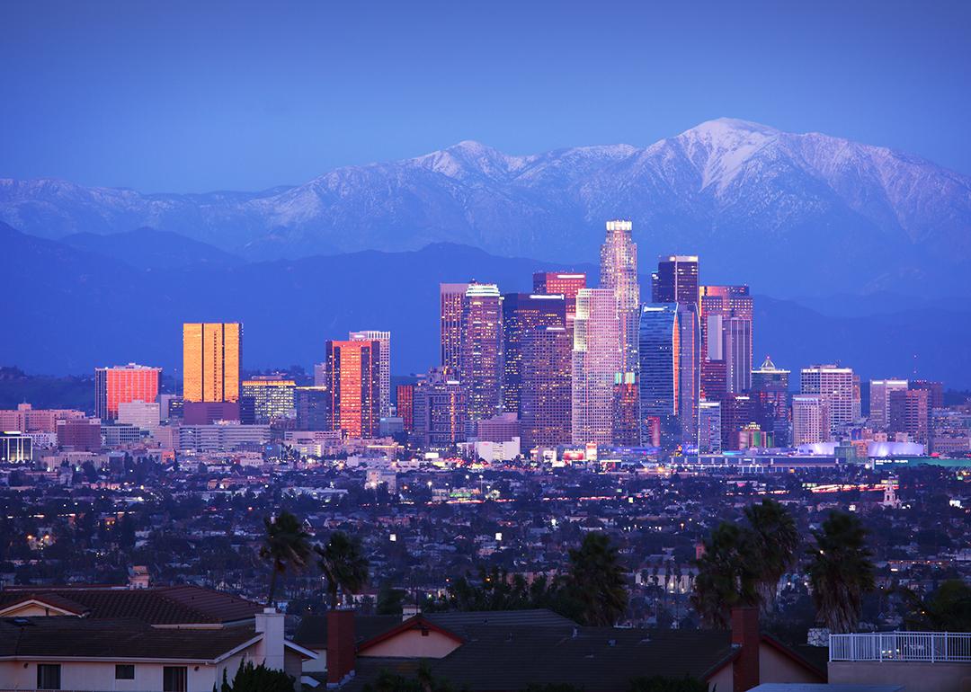 A view of downtown Los Angeles skyscrapers over snowy mountains during twilight.