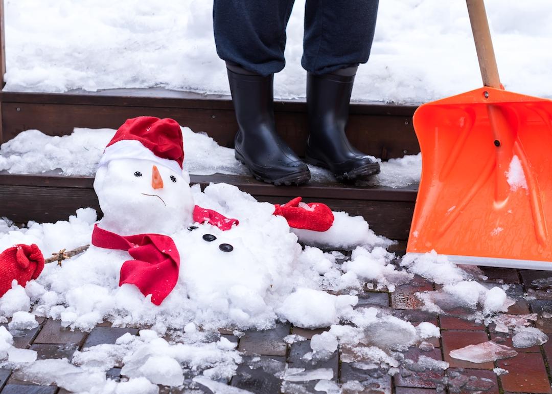 A snowman in red mittens, a scarf, and cap is melting outdoors on wet pavement next to a person's boots and shovel.