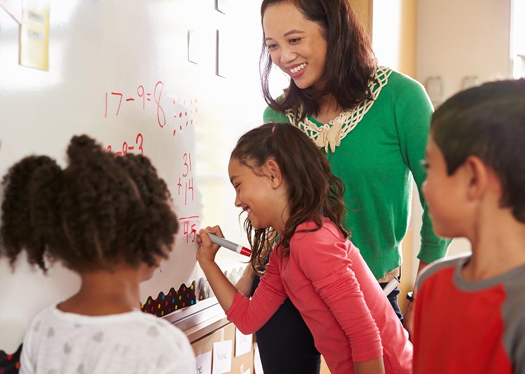 A grade school teacher happily looks over pupils answering a math exercise on the board.