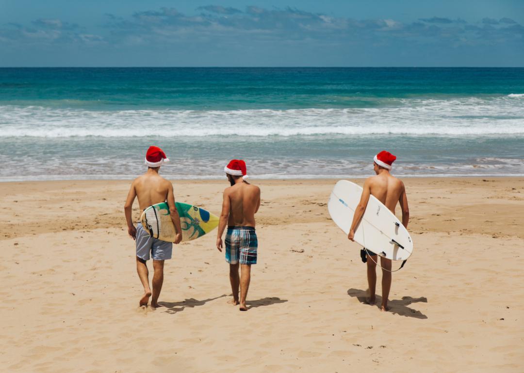 Three people walking on the beach holding their surfboards and wearing Santa hats.