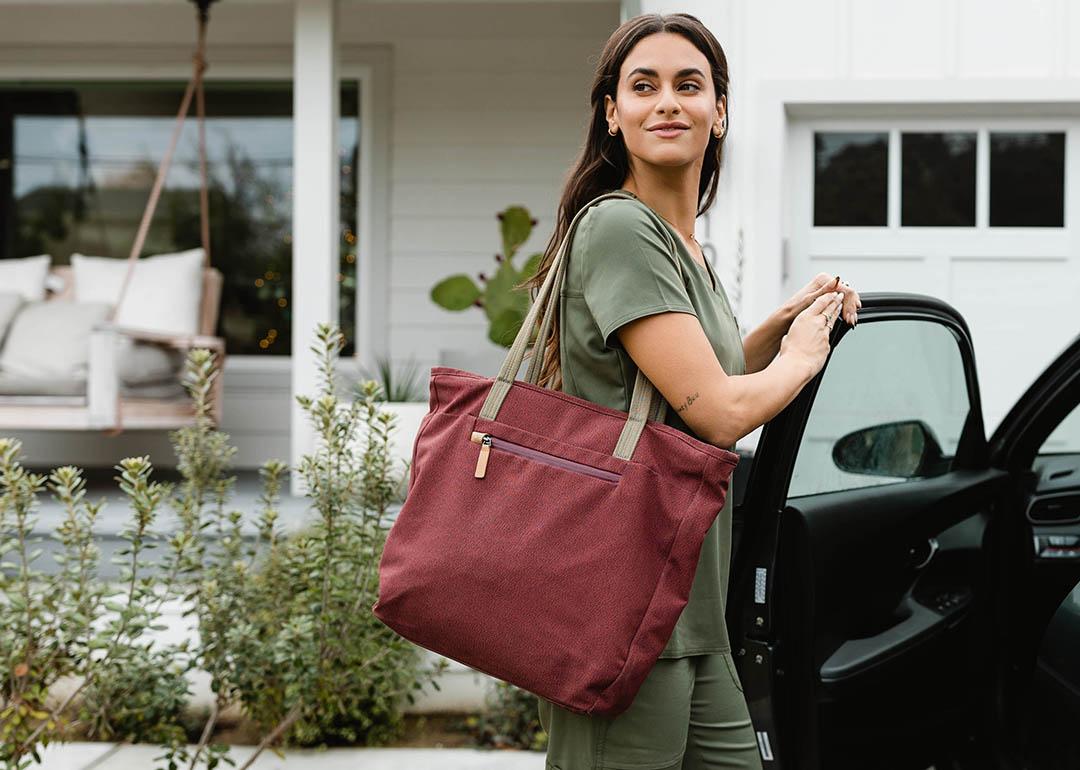 Travel nurse wearing olive green scrubs and carrying a tote bag looks over her shoulder with one hand on an open car door parked in driveway.