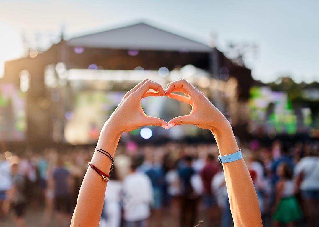 Concertgoer holds hands up to form a heart shape in the foreground of an outdoor music festival with stage in background.