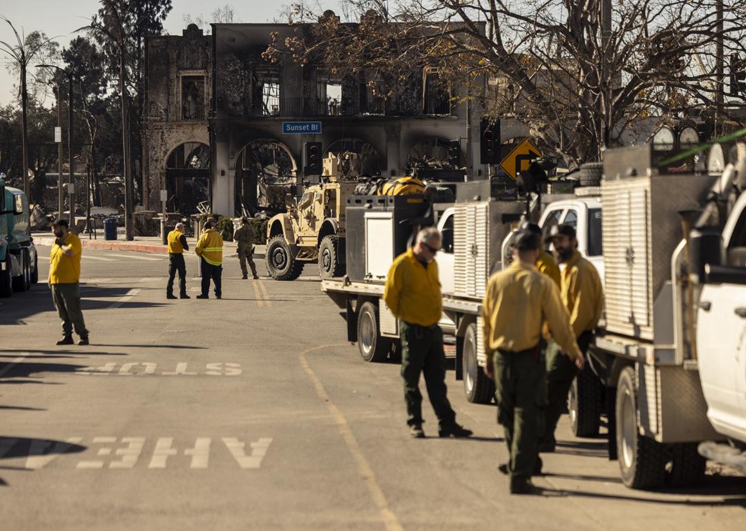 A charred building is seen in the distance as private firefighting company employees, hired to protect Rick Caruso's Palisades Village mall from the Palisades Fire, gather near their vehicles.