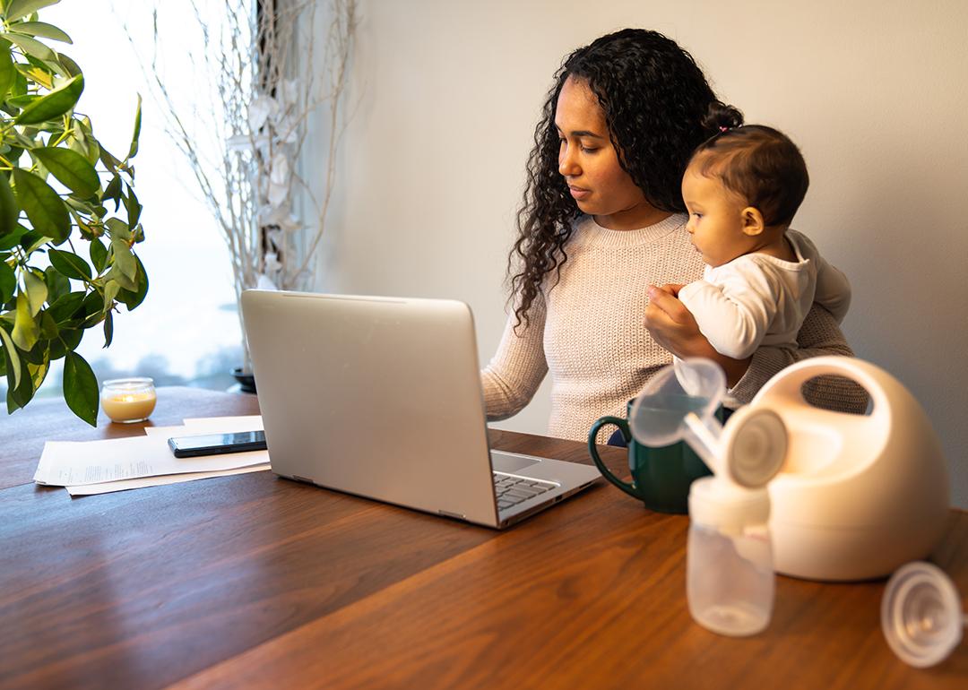 An African-American mother holds her baby daughter while taking notes and using a laptop.