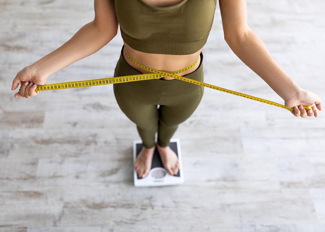 A woman on checking body measurements with a tape and checking weight on a scale.