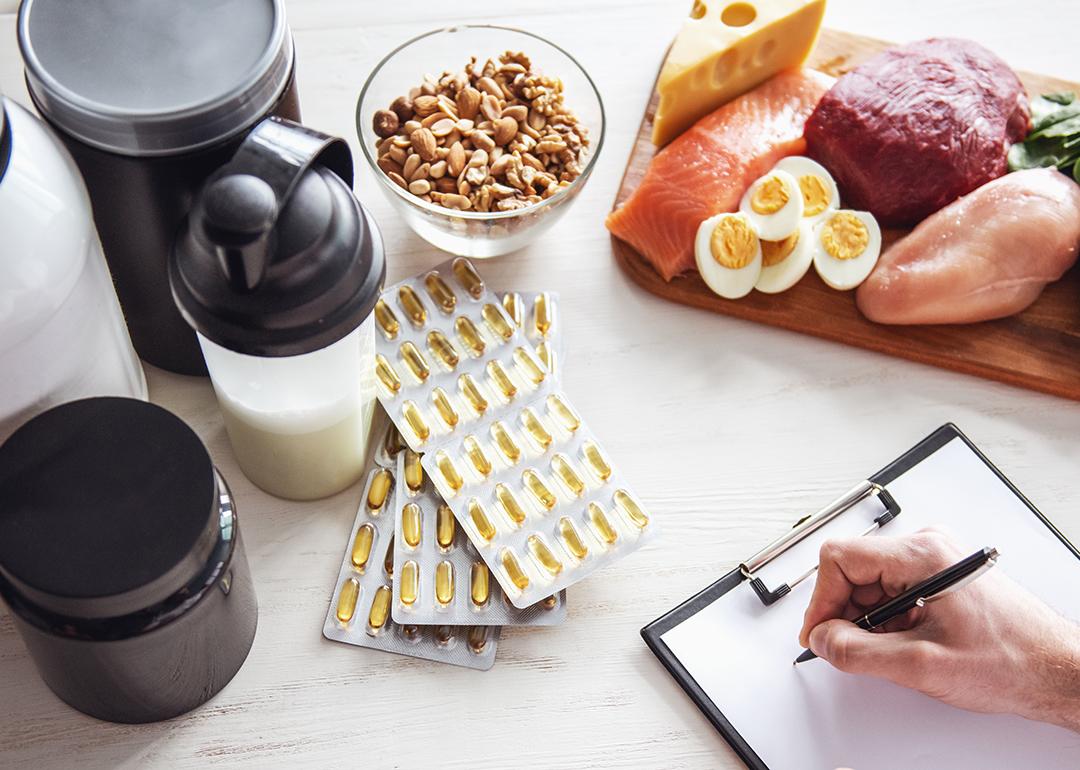 A set of food supplements such as protein powder and fish oil supplements on a table with fresh food while a person writes a diet plan.