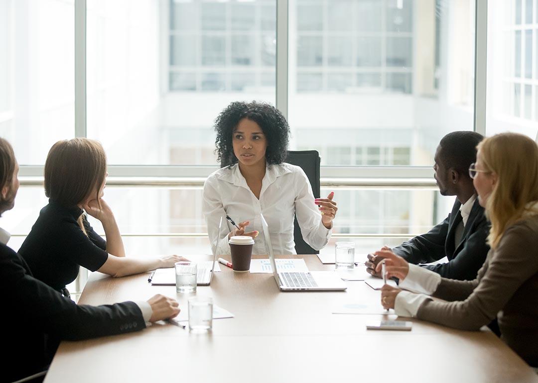 Black woman at head of table leads discussion with colleagues on each side of table.