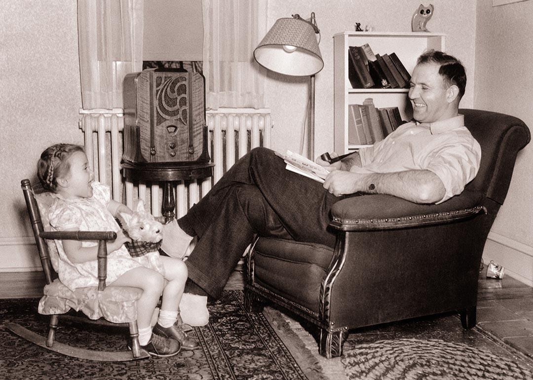 1930s Smiling father sitting in overstuffed chair watching daughter in small rocking chair both listening to wood cabinet radio.