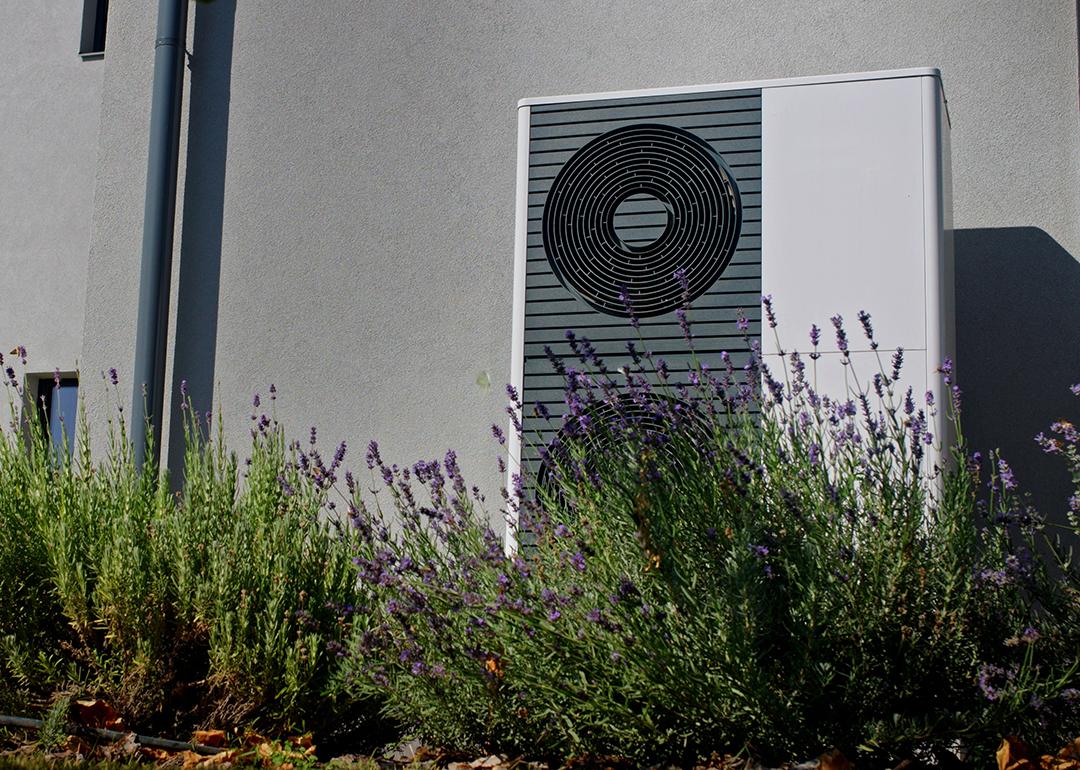 A modern outdoor heat pump mounted on a wall of a home surrounded by lavender plants.