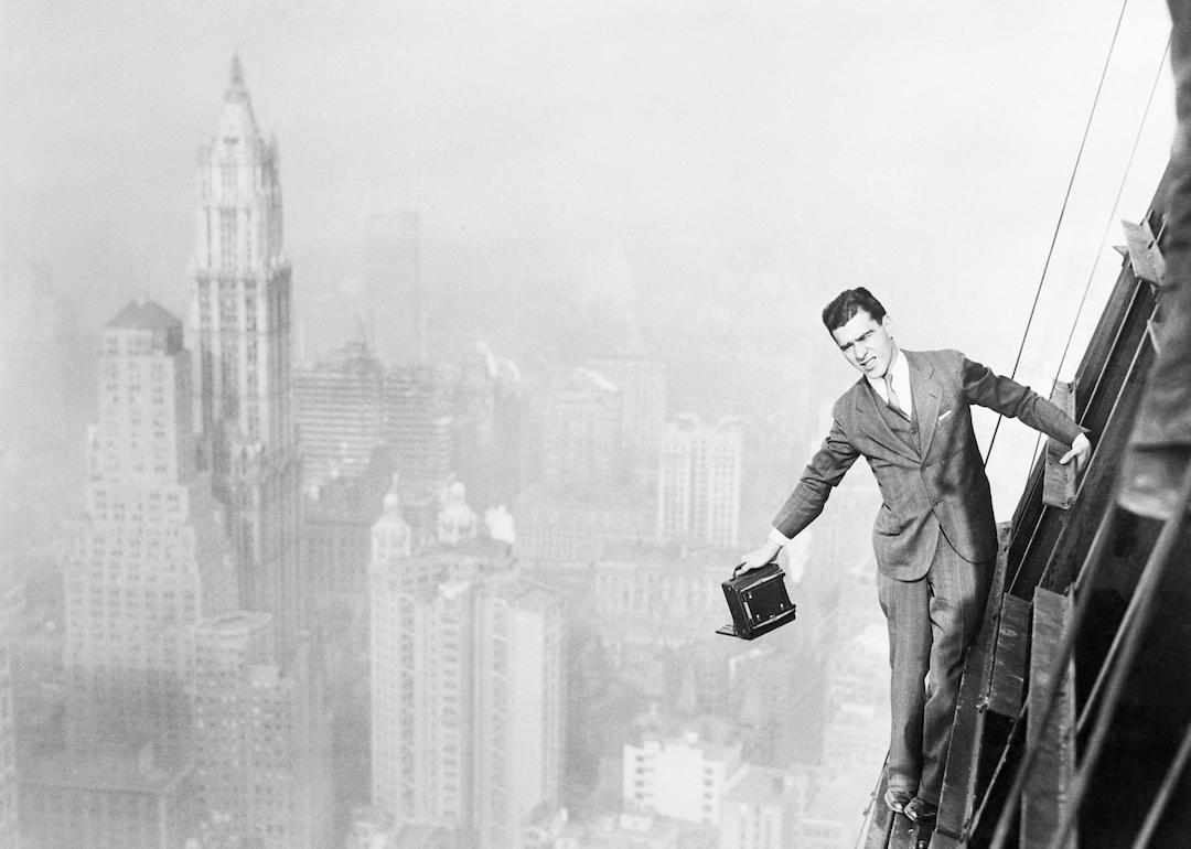 Photographer Jack Reilly precariously perched from the 72nd story of the world's tallest building, the Bank of Manhattan structure, in progress of construction in Wall Street, New York City in 1929. A panorama of New York's prominent business section is seen in the background, with the Woolworth building, in the upper left.