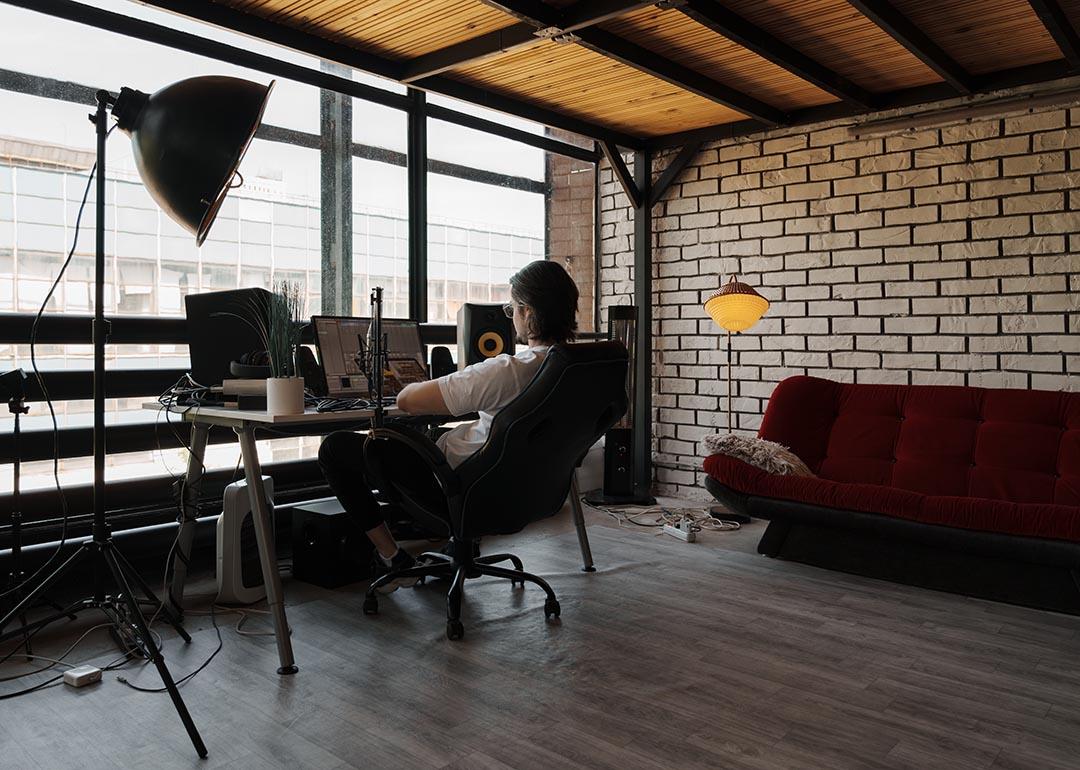 Man working on computer in a modern industrial loft office with large windows, minimalist furniture, and comfortable ambiance.