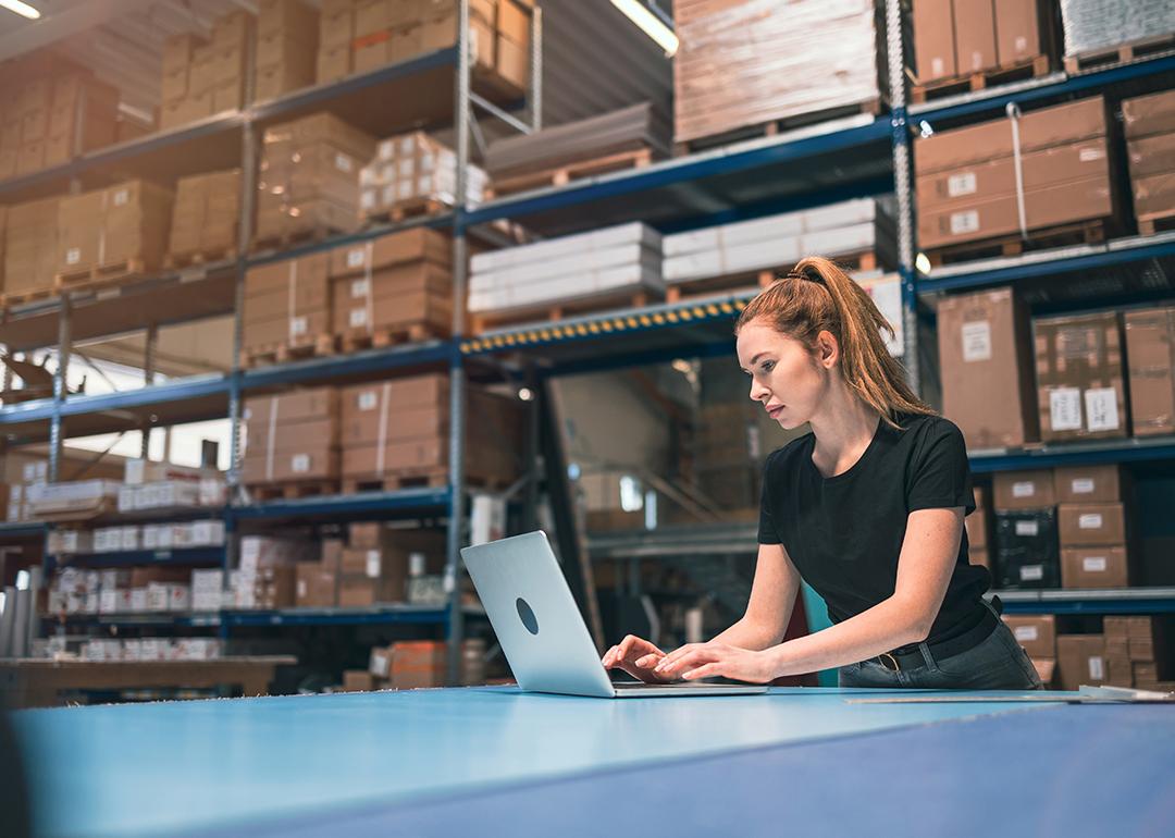 A woman managing logistics and using a laptop in a warehouse.